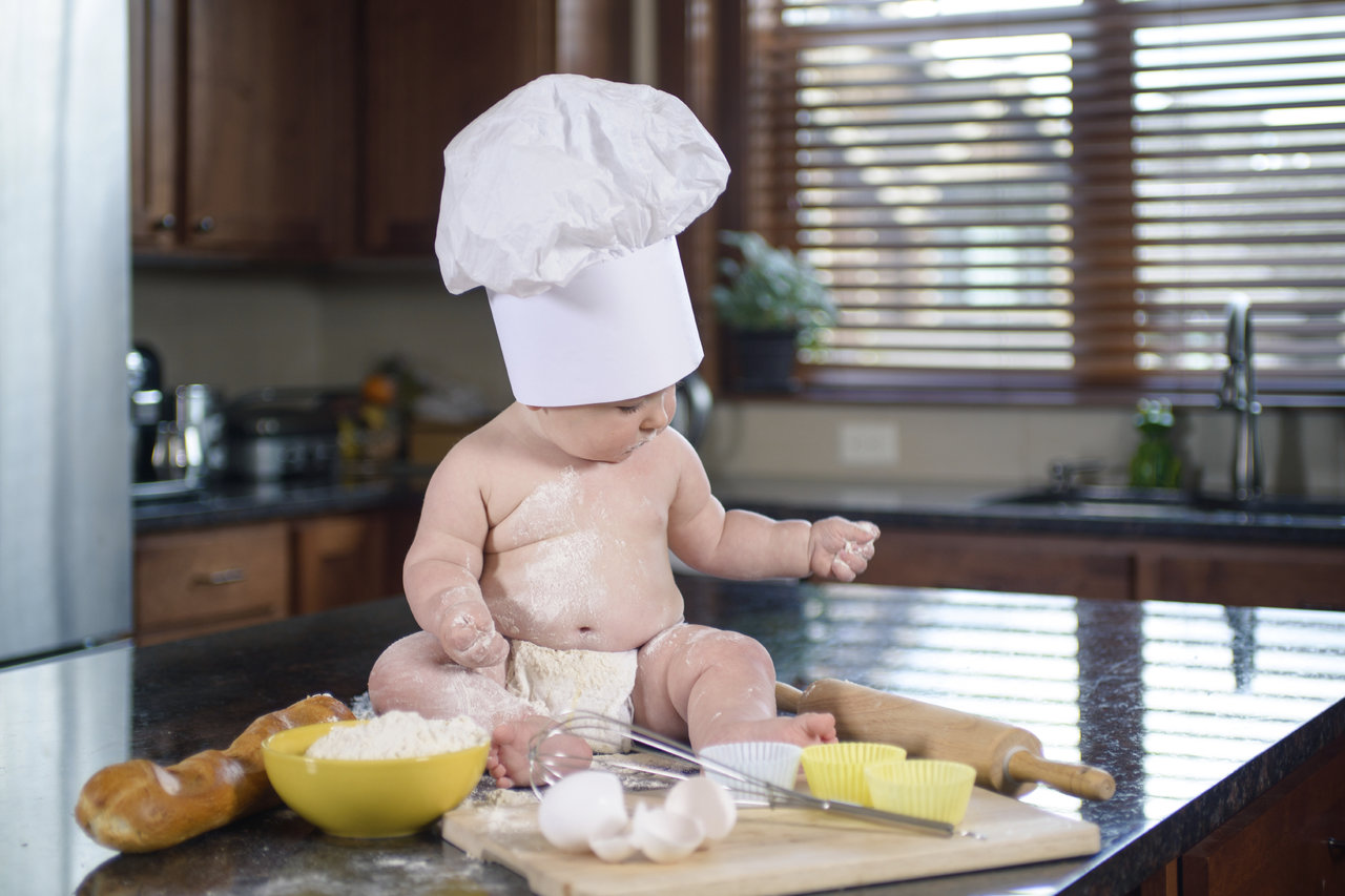 Baby boy in chef hat covered with flour sitting on kitchen table