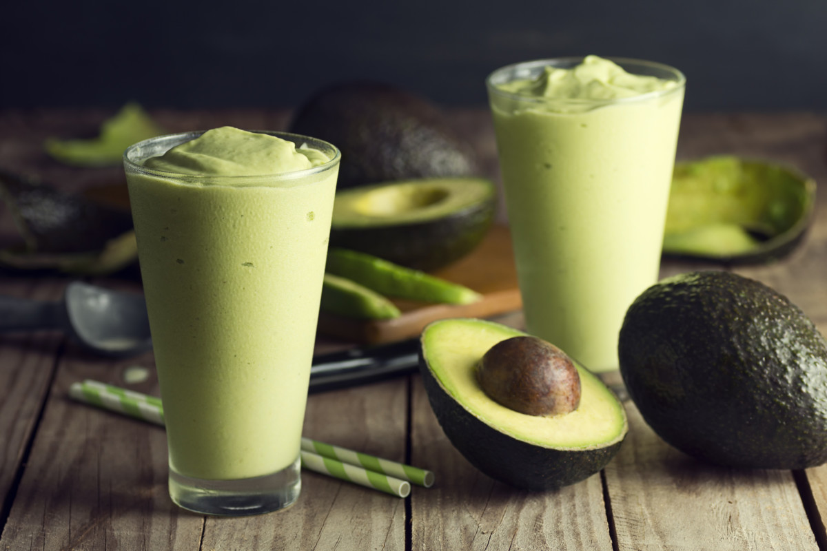 A freshly made avocado shake, or smoothie, in glasses on a wooden table. Avocados, a knife, a cutting board and an ice cream scoop are in the background. Avocado shakes originated in southeast Asia and are also found in Brazil, but are becoming popular across the globe.