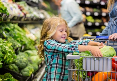 Elementary age Caucasian blonde little girl is smiling while placing lettuce in shopping cart. Child is shopping for produce and healthy food with her mother in local grocery store. Child is wearing a plaid shirt. Other customers are shopping in background.